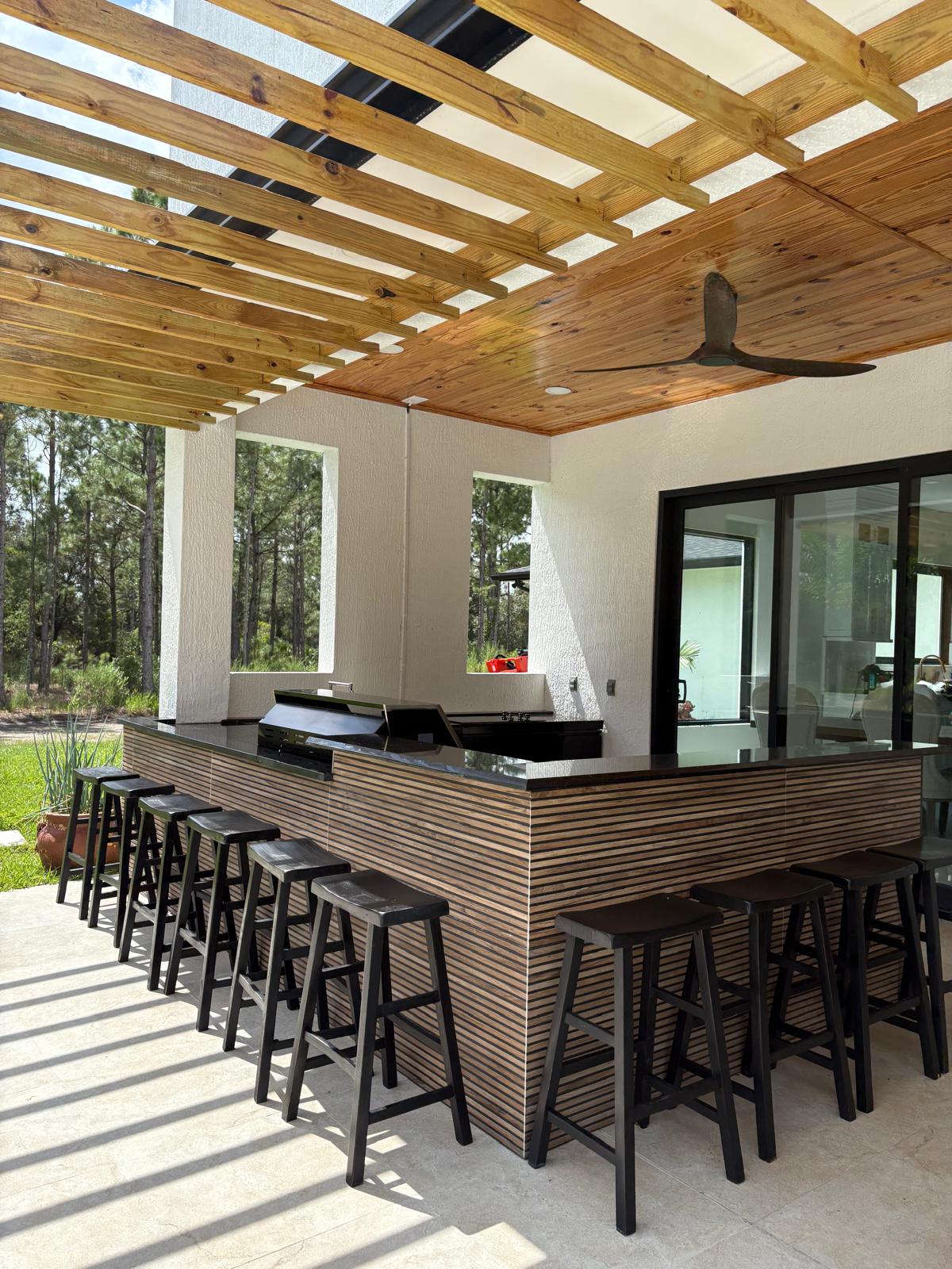 Modern U-shaped outdoor kitchen under pergola with dark cabinets and black granite counters